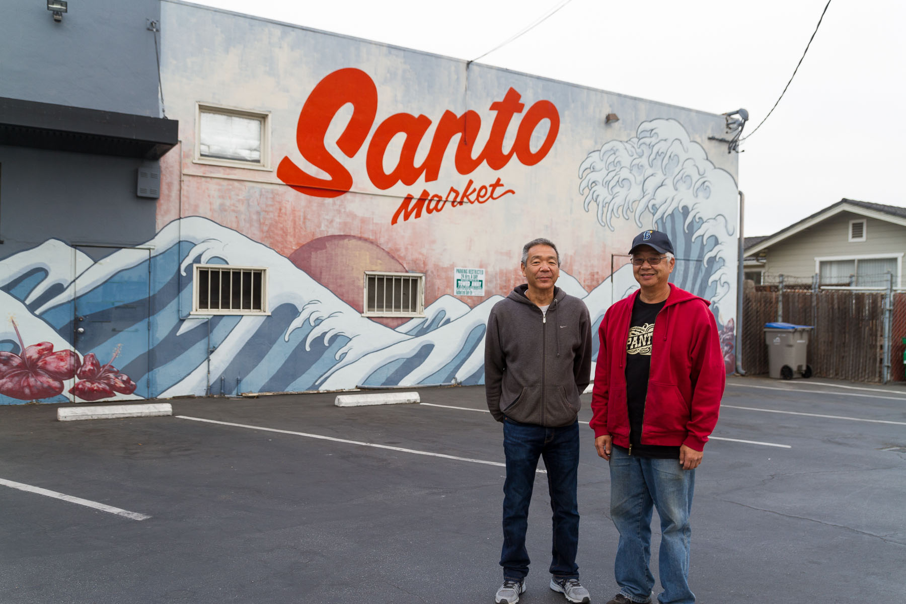 Two men stand in front of a large, ocean-themed mural on the exterior wall of Santo Market.
