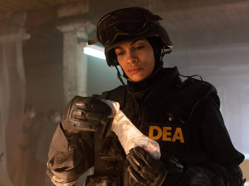 A woman wearing a SWAT uniform and helmet, as well as a vest marked DEA, frowns as she examines a plastic bag containing pills.