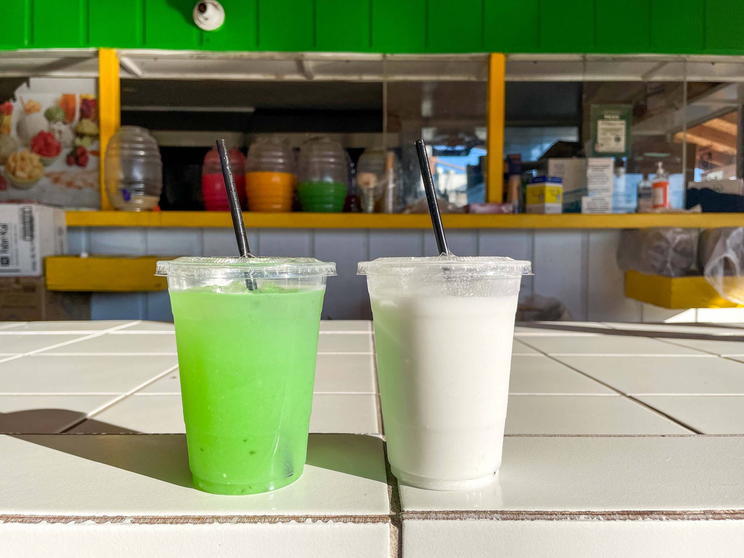 Two agua frescas, one green and one white, arranged on a table in front of a juice stand.
