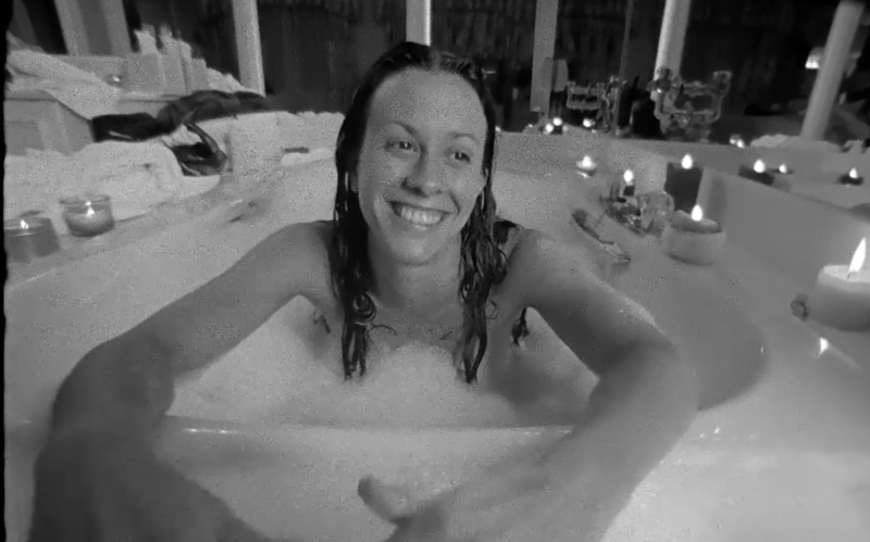 A black and white image of Alanis Morissette sitting in a bubble bath smiling broadly, long hair wet from the water. A variety of candles burn in the background.