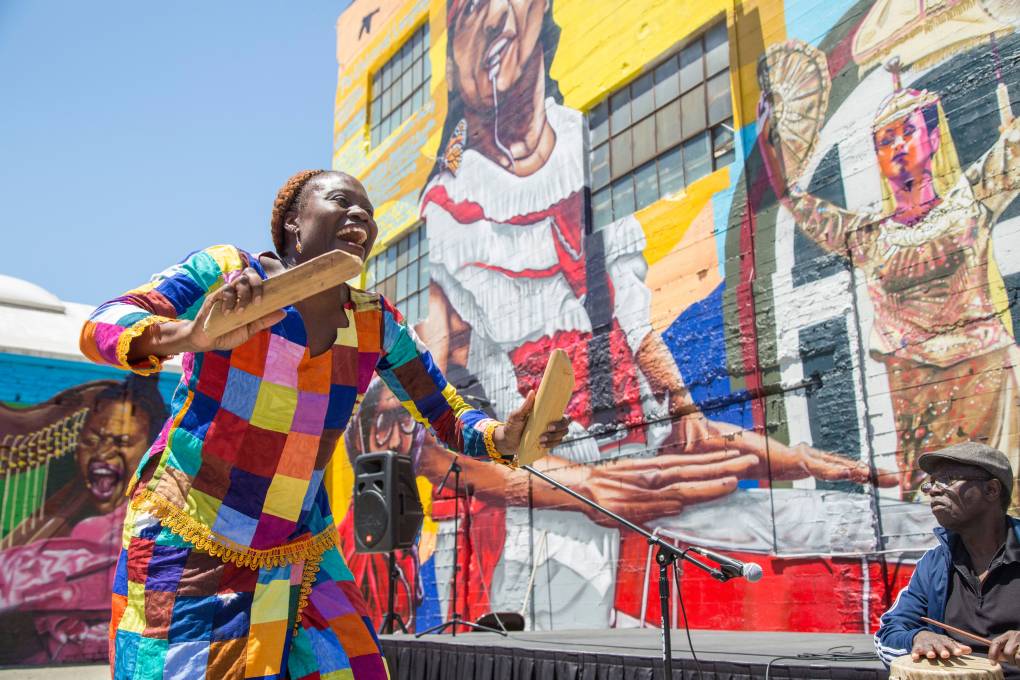 Esailama Artry-Diouf from the West African group Diamano Coura is dressed in bright multicolored fabric, as she dances in front of the now invisible mural in downtown Oakland.
