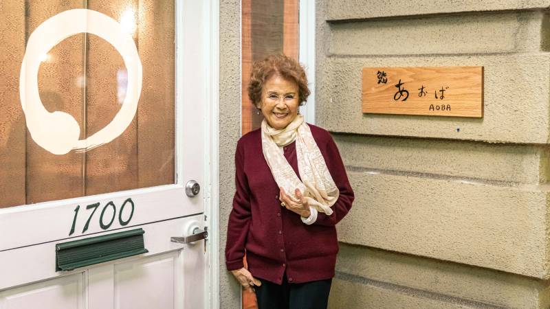 An older woman in a white scarf stands next to the entrance to Aoba restaurant.