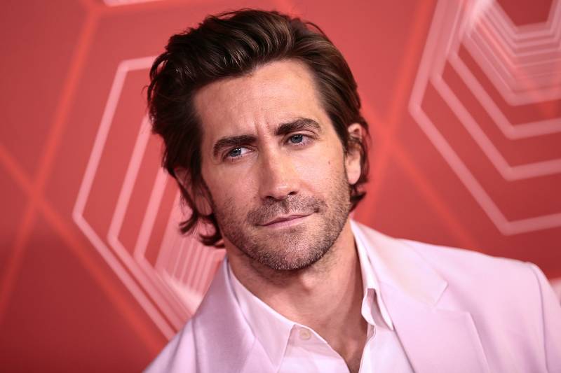 Jake Gyllenhaal wears a pale pink shirt and suit jacket in front of a red and pink wall.