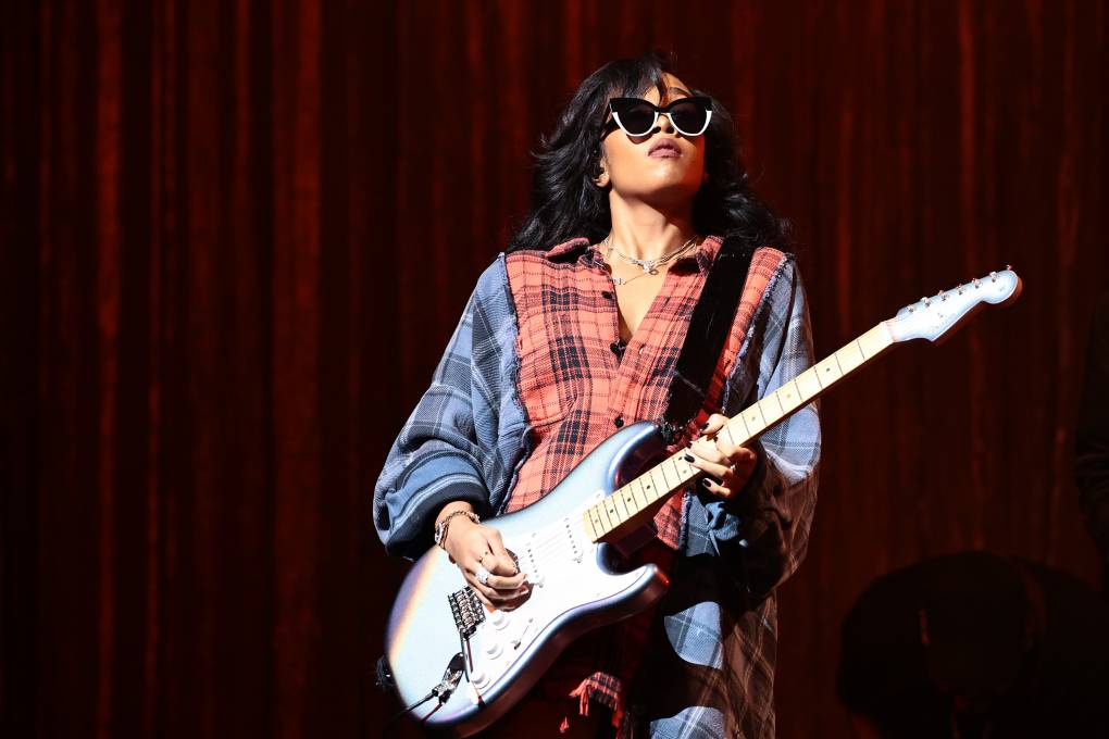 A Black woman wearing an oversized plaid shirt and sunglasses passionately plays an electric guitar on stage.