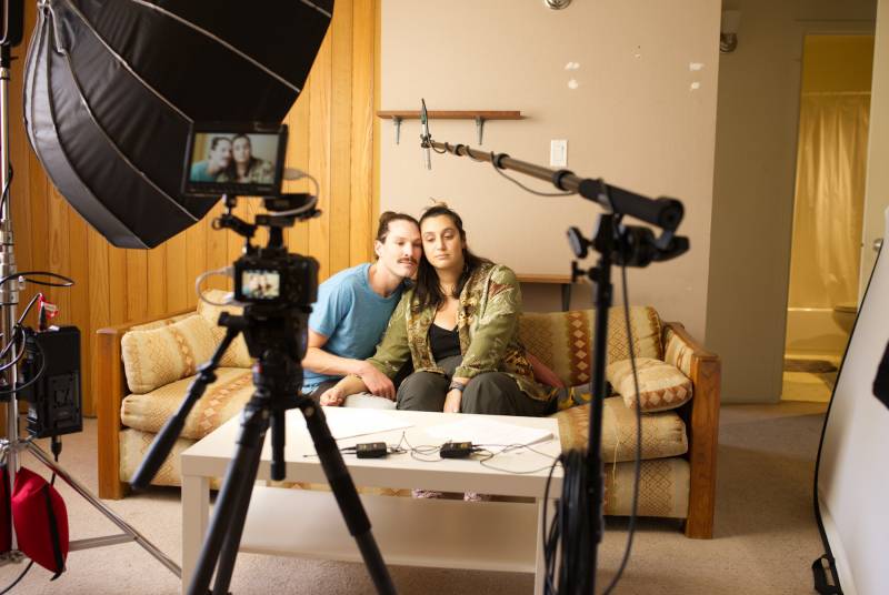 A couple sits on a loveseat, looking forlorn, surrounded by camera equipment
