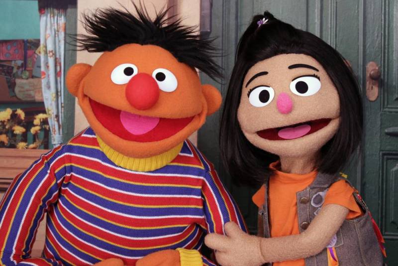 The muppet Ernia, in a striped shirt, poses with the new muppet Ji-Young, in an orange shirt and vest.