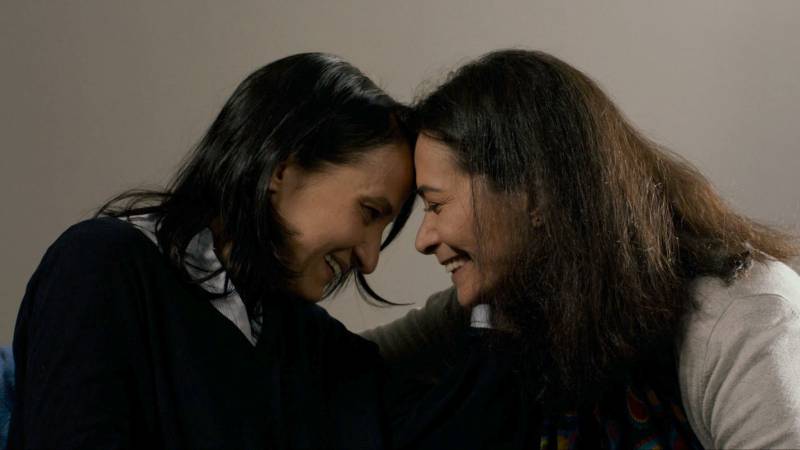 Two women lean toward each other, smiling and touch foreheads.