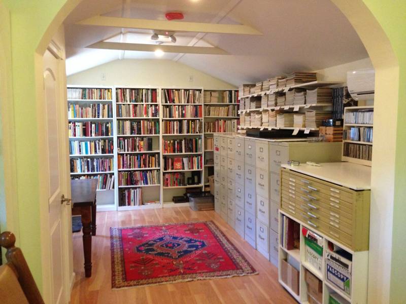 A door opens to flat files and a wall of bookshelves with a red rug on the floor.