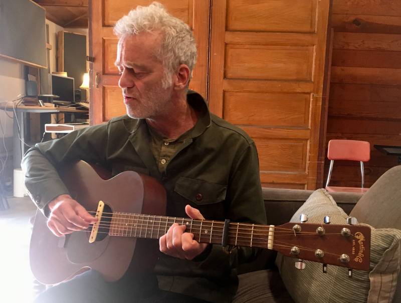 A man with short grey hair and beard plays a vintage acoustic guitar in a wood-paneled room as sun shines in from the left.