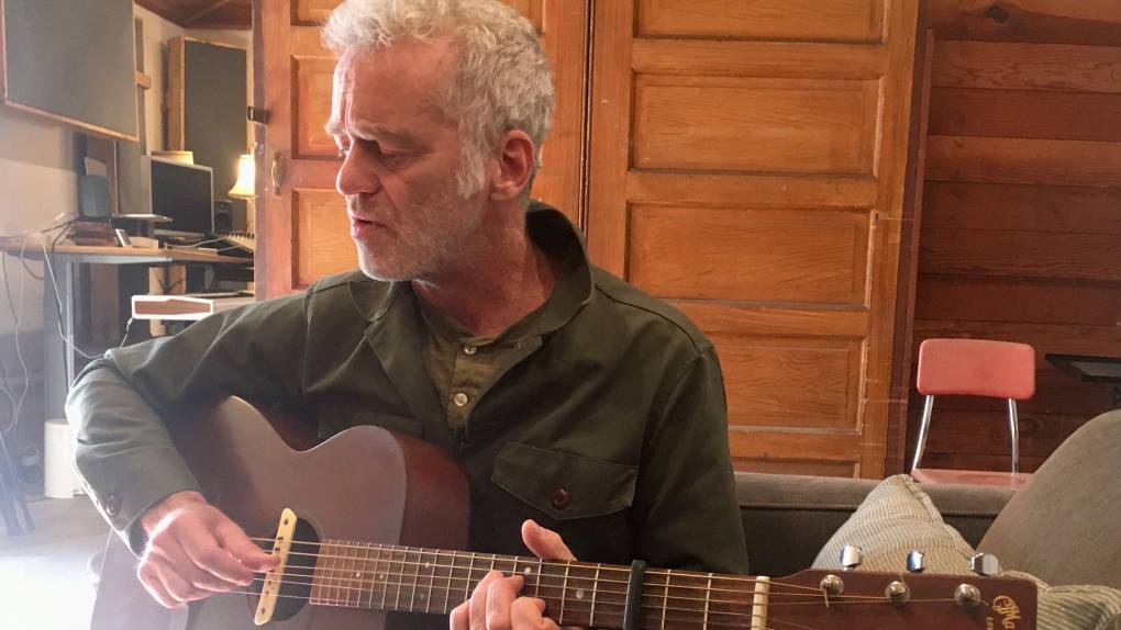 A man with short grey hair and beard plays a vintage acoustic guitar in a wood-paneled room as sun shines in from the left.