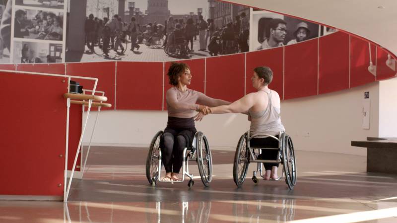 Disabled dancers Alice Sheppard and Laurel Lawson dance, holding hands as they face each other in their chairs. 