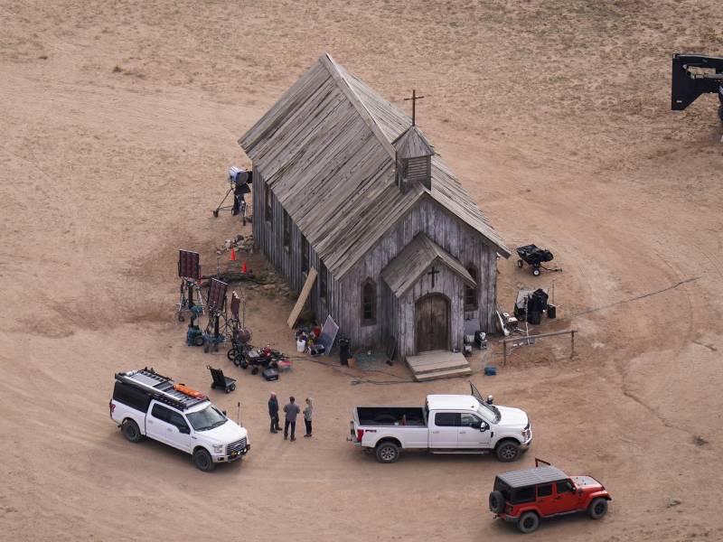 An aerial shot of an old wooden church in the desert. Two modern trucks and a car are parked outside.