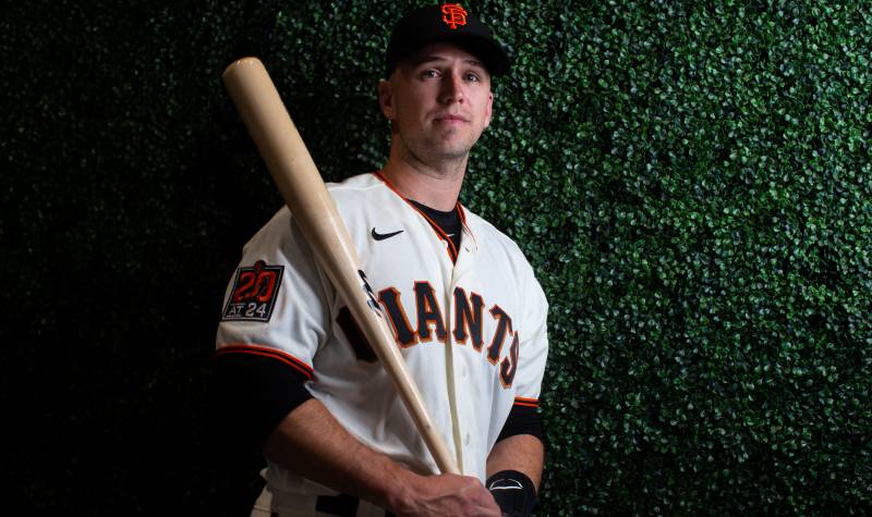 A white man in a San Francisco Giants uniform stands against a green backdrop, casually clutching a baseball bat.
