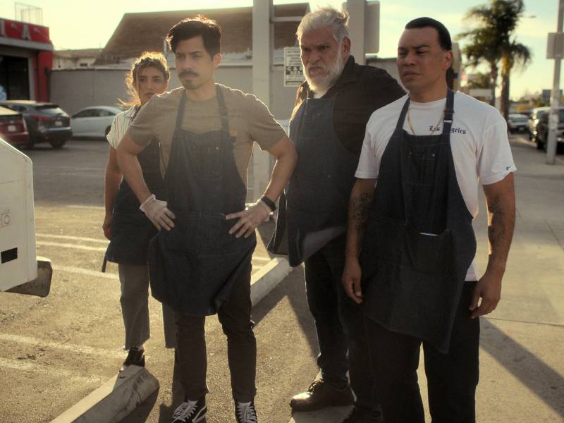 An older Latino man stands in a parking lot with two younger men either side of him wearing aprons, and a young woman, also wearing an apron, to the left. They all look concerned.