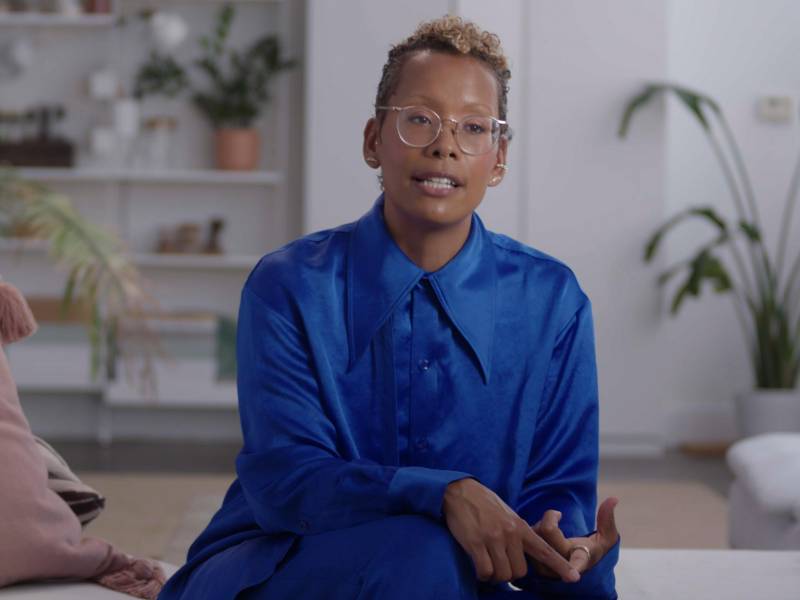A Black woman with cropped hair and glasses wears a blue silk shirt and sits talking in a white room with plants visible behind her.