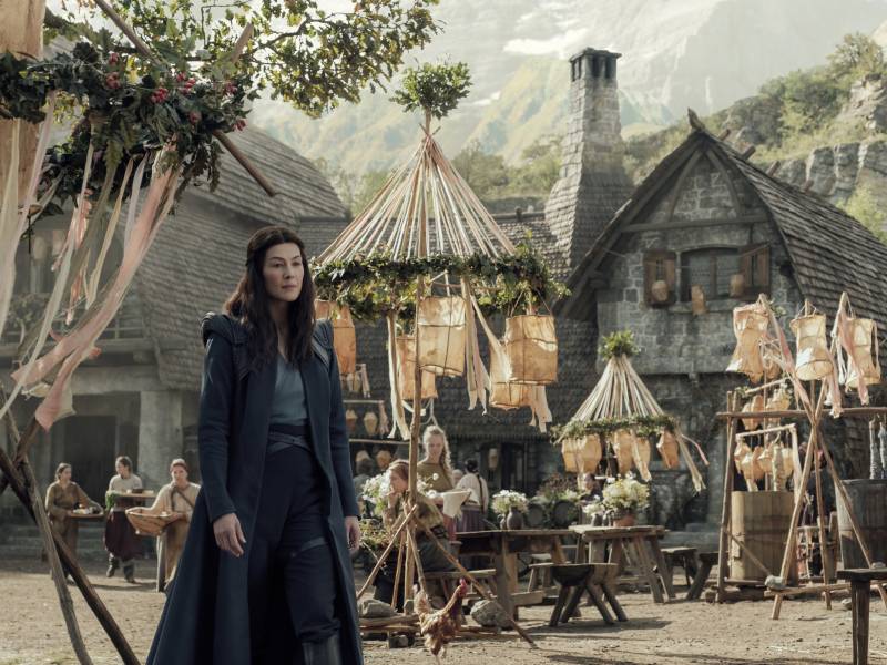A woman in dark blue, futuristic looking long coat, pants and top stands before a village that more closely resembles Medieval times.