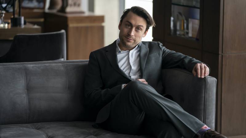 Kieran Culkin sits on a grey couch in character as Roman Roy. He is wearing a grey suit and white shirt with no tie.