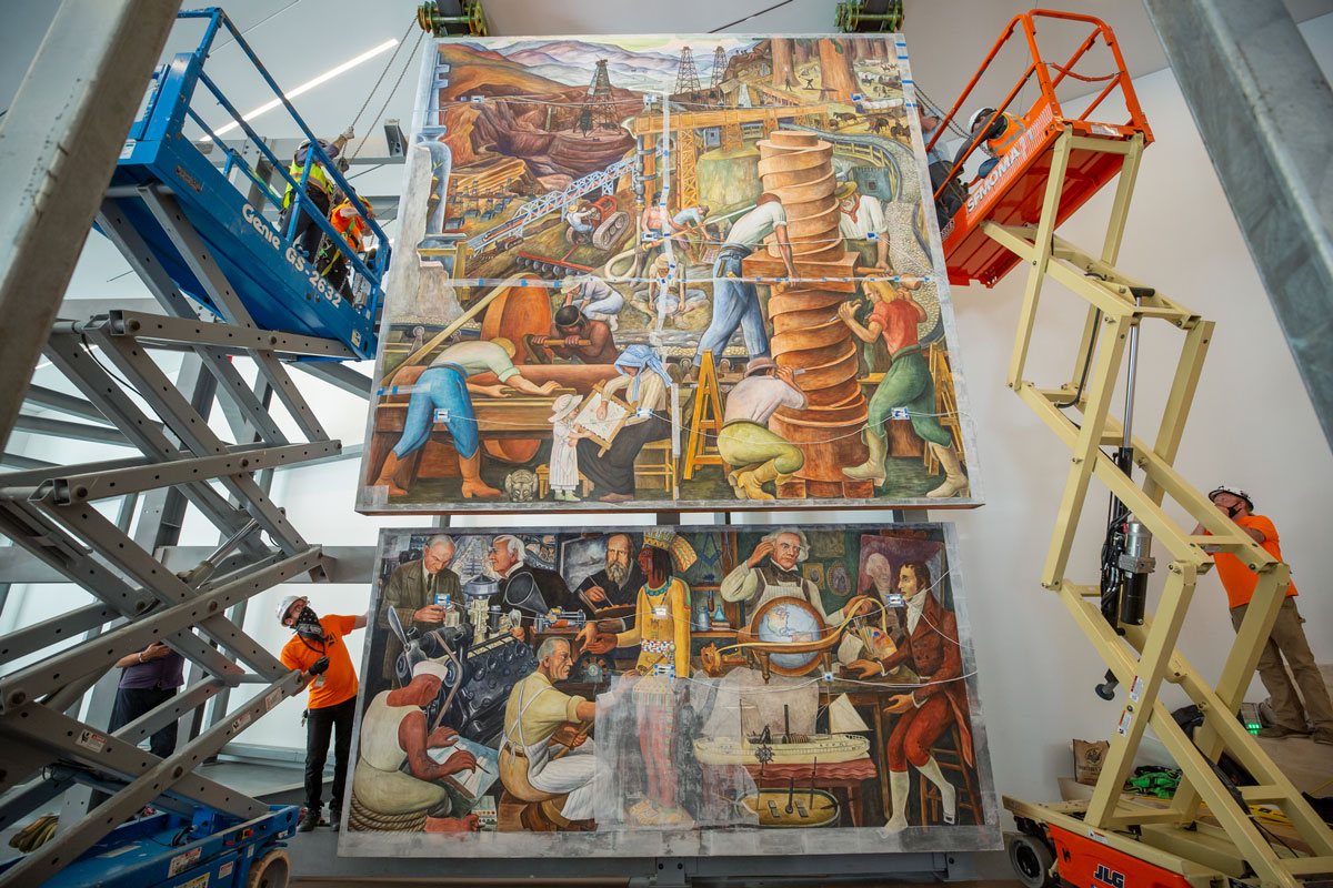 Two scissor lifts and workers flank two vibrant panels of fresco painting.