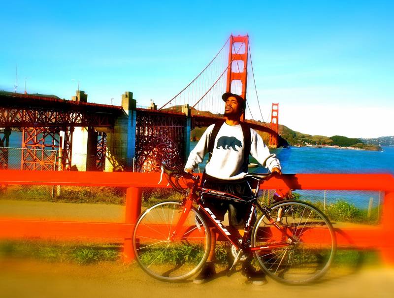 Pendarvis Harshaw in front of the Golden Gate Bridge holding his bike and looking off in the distance.
