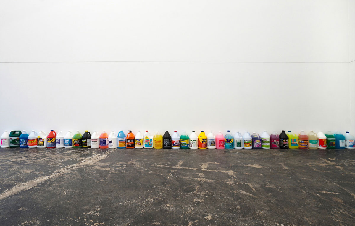 A row of multicolored gallon jugs in a row against a white wall.