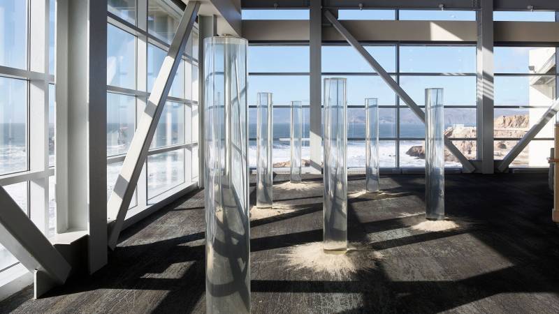 Tall clear cylinders contain ing water in a room of windows overlooking the ocean.