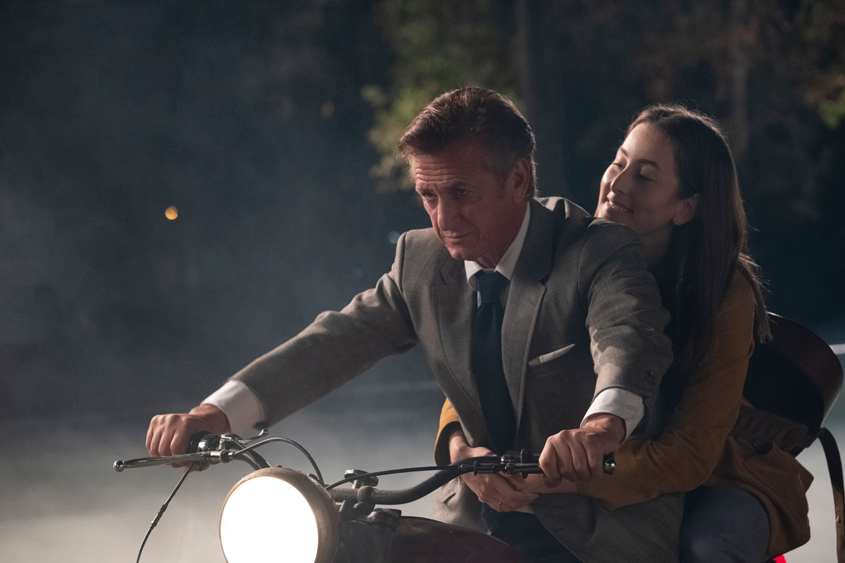 Older man in gray suit drives a motorcycle with young woman behind him.