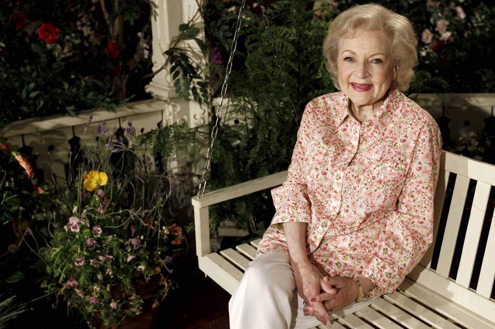 Betty White smiles, sitting on a white porch swing next to garden flowers.