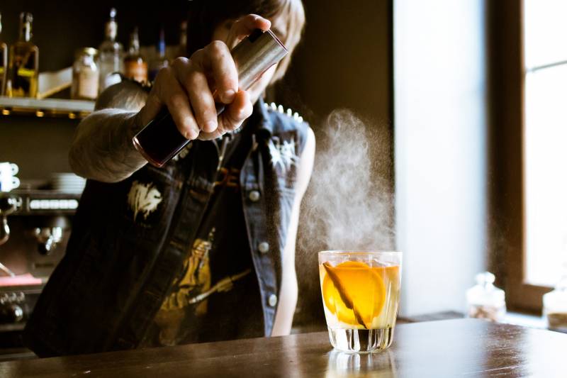 A bartender wearing black sleeveless denim jacket pours out a cocktail.