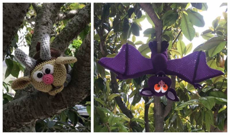 Left: A crocheted brown goat with a round pink nose peaks over a tree branch. Right: A purple and black bat with fangs hangs upside down from a tree branch.