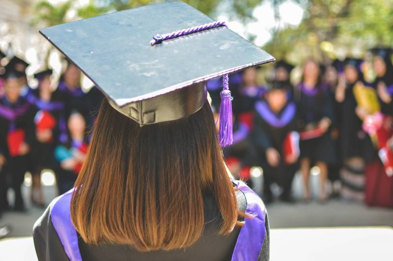 A woman wearing a cap and gown, viewed from behind, looking onwards to her graduating classmates.