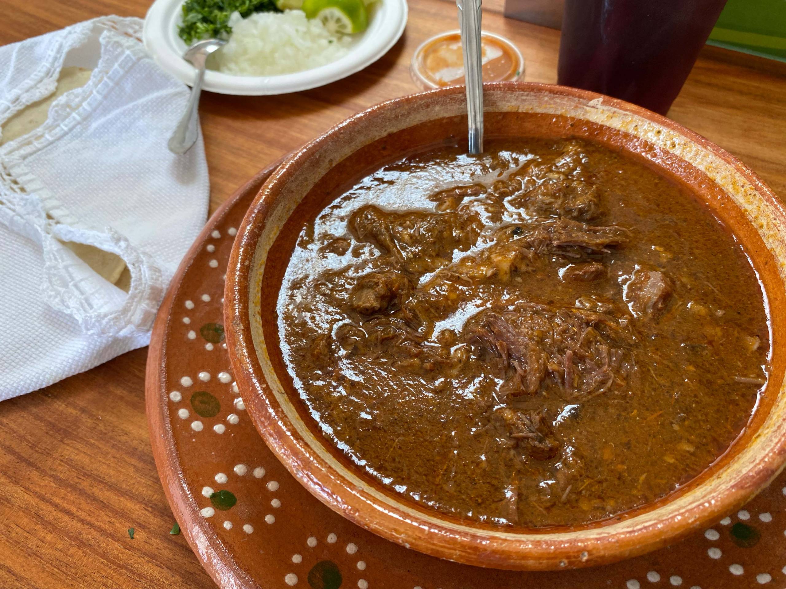 An earthenware bowl of beef barbacoa, with tortillas wrapped in a dish towel on the side.