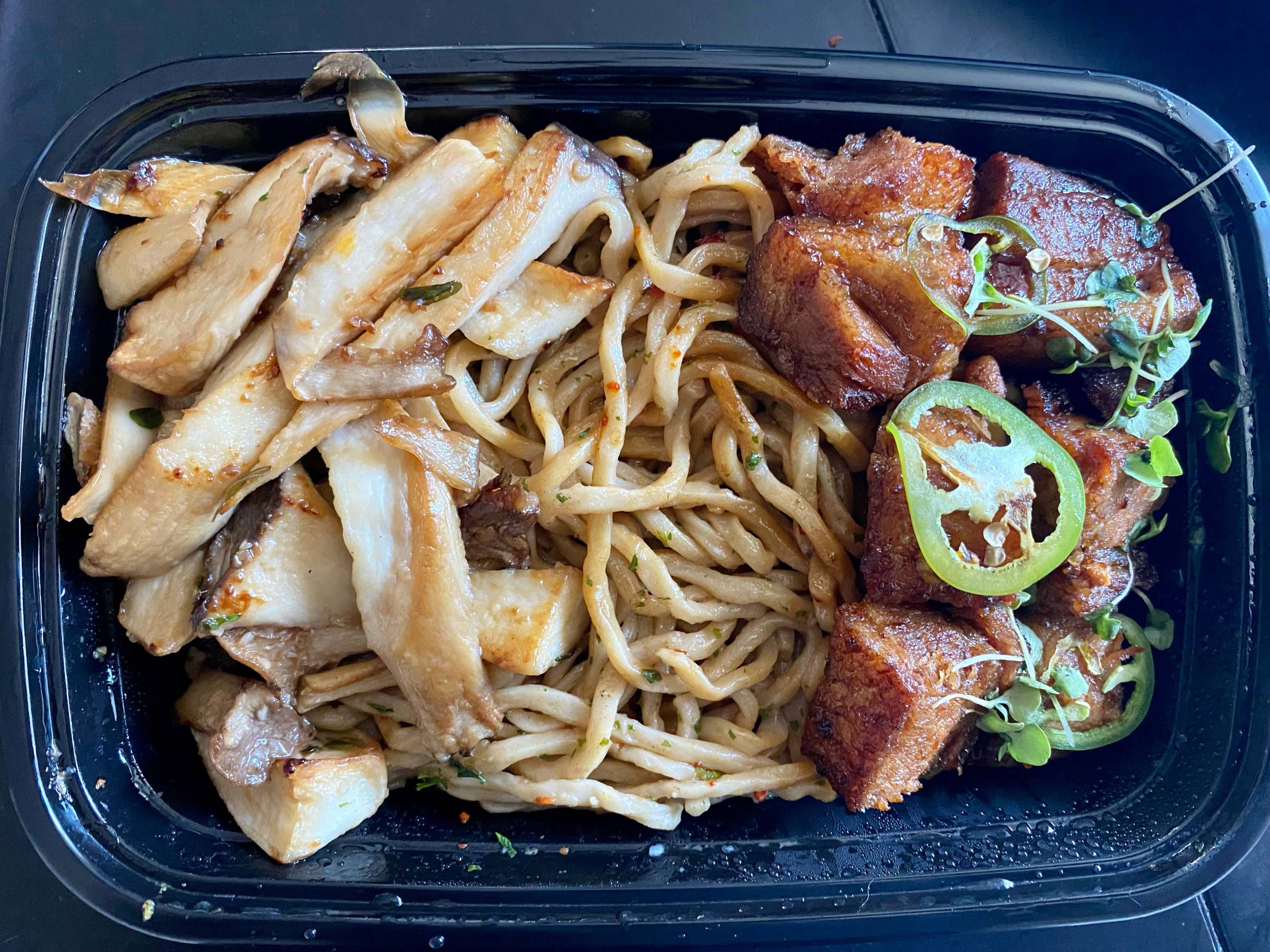 Overhead view of a takeout container of garlic noodles, roasted mushrooms and crispy pork belly.