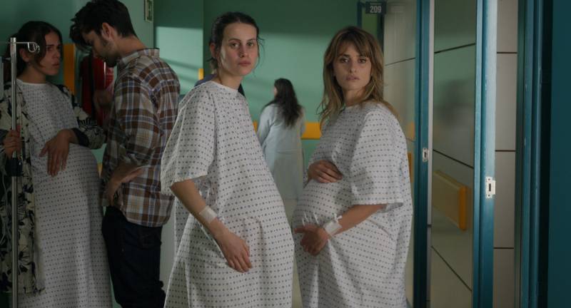 Two pregnant women in hospital gowns stand with their bellies nearly touching.