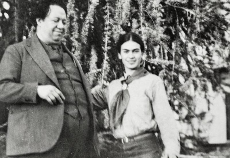 A black and white photo shows Diego Rivera on the left and and a smiling Frida Kahlo on the right posing in front of a tree at the home of Luther Burbank.