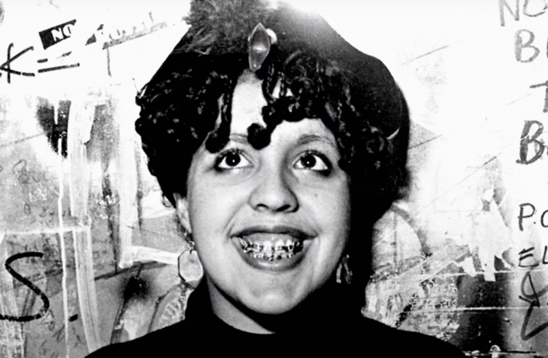 A black and white image of a Black woman wearing her hair in short tight curls that frame her face, grinning broadly with braces on her teeth. Graffiti covers the wall behind her.