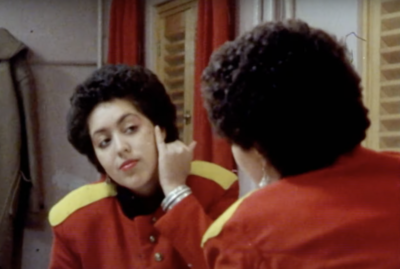 The singer for X-Ray Spex, wearing a red and yellow band jacket, gazes into a mirror and applies make-up to her cheeks.
