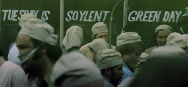 A crowd of people, all wearing beige bonnets, gather in front of shop windows painted with the words, "Tuesday is soylent green day."