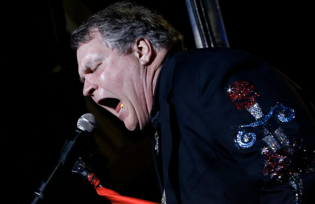 Meat Loaf, wearing a black suit on stage and sweating under the lights, sings passionately into a microphone.