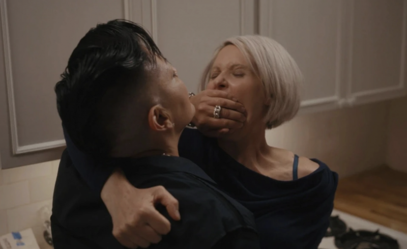A woman with a partially shaved head embraces another woman with a grey bob. The first woman covers the mouth of the second woman during a passionate embrace. They are standing near a stove.