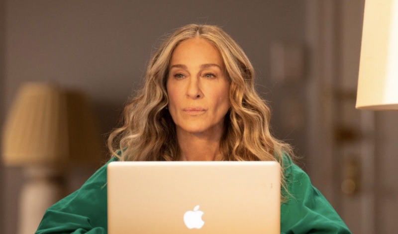 A woman with long wavy blonde hair sits in a window, wearing a green shirt, behind an open Apple laptop, looking thoughtfully out to the horizon.