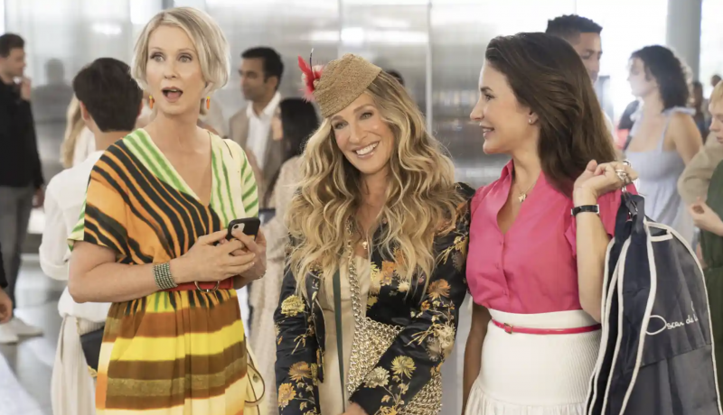 Three women in colorful, stylist outfits stand together in a line, one shocked, one smiling, one distracted.