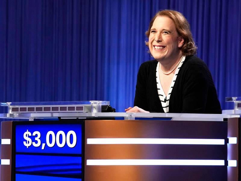 A woman smiles broadly from behind a game show podium, $30,000 written on the screen in front of her. She wears a black sweater and pearl necklace with matching earrings. Behind her hangs a dark blue curtain.