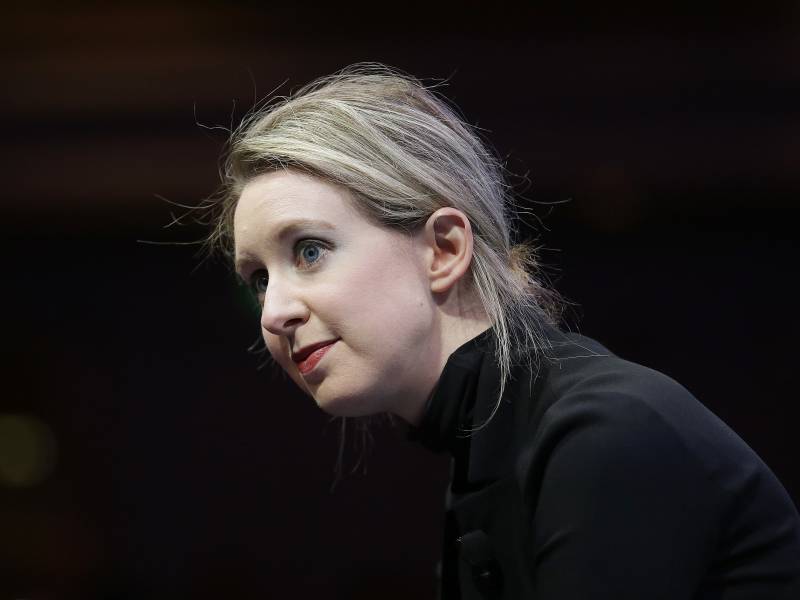 Elizabeth Holmes, in her trademark black turtleneck, blonde hair pulled back into a messy ponytail, leans forward to listen to someone.
