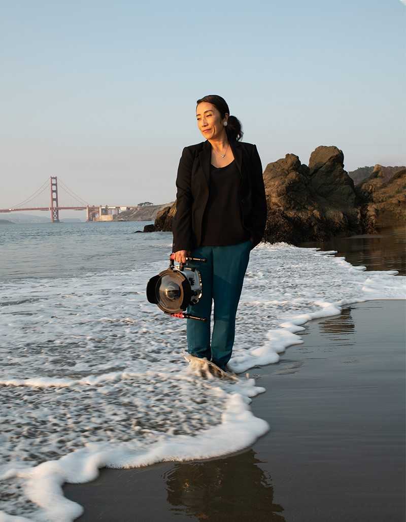 Erena Shimoda holds a large camera, as she stands on a beach as the tide comes in with the Golden Gate Bridge in the background.