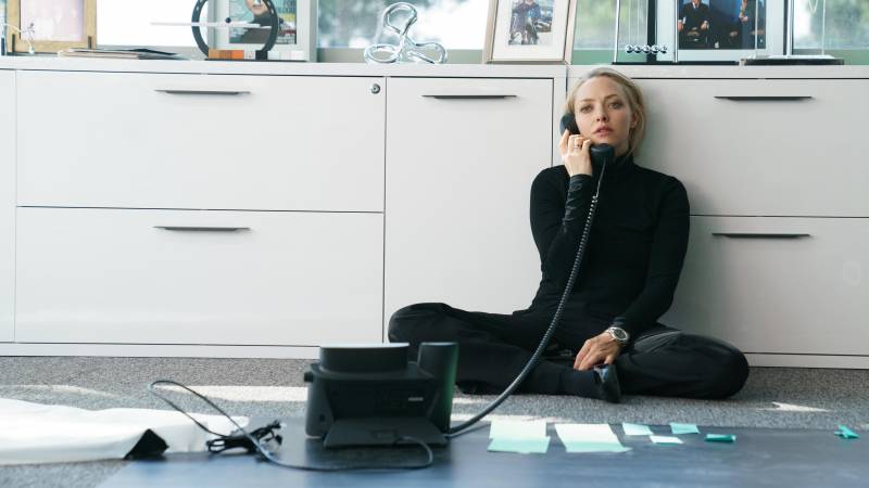 Amanda Seyfried, in character as Elizabeth Holmes, wears black pants and a black turtleneck and sits on the floor talking on the phone, her back against white filing cabinets, papers spread out before her.