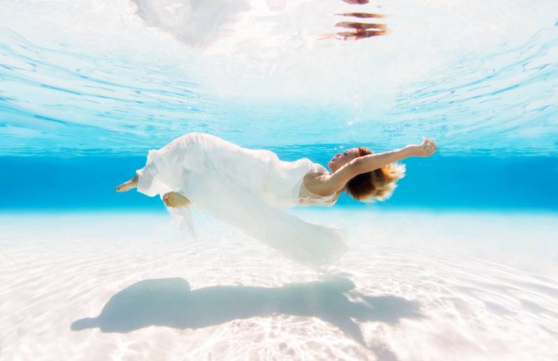 a woman floats horizontally in the pool, it's bright, she wears a white dress with her arms stretched above her head. 