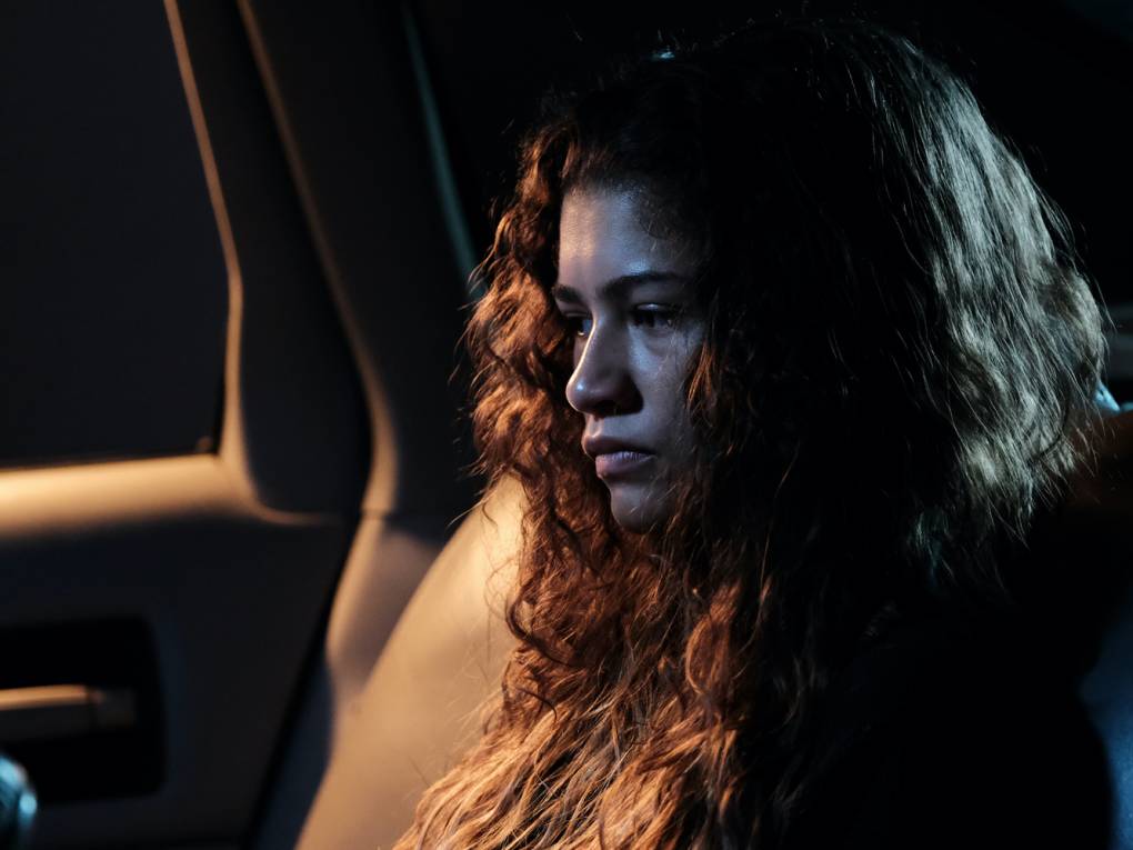 A young woman sits in the back of a car, looking stunned and numb. It is dark outside.