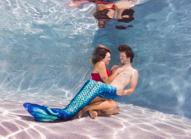 Underwater we see a man in a blue mermaid tail smiles at a woman holding him in a red swimsuit.