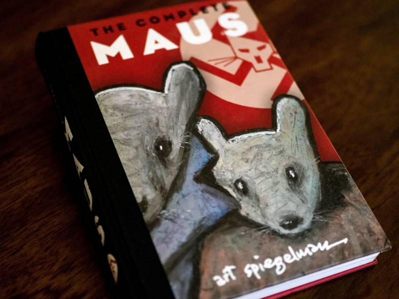 A photo of the cover of graphic novel, 'Maus' featuring a big mouse and a small mouse, wearing human coats in front of a red wall featuring Nazi symbolism.