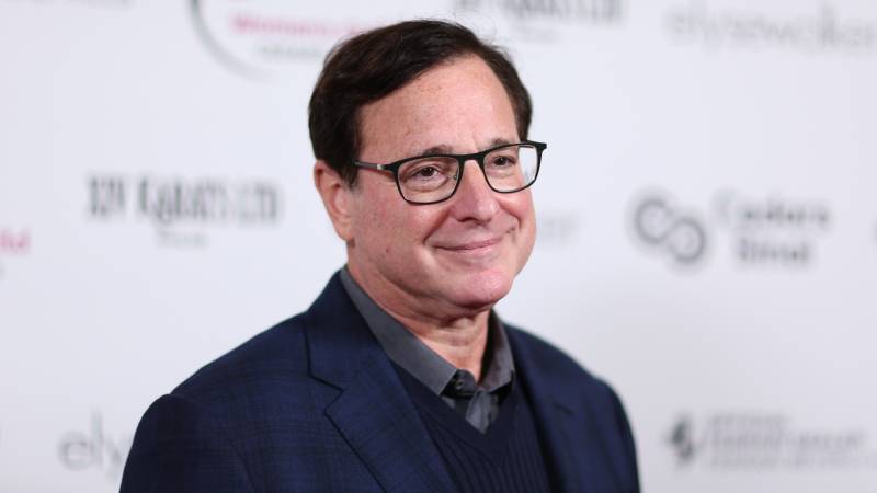Bob Saget, wearing a casual blue suit smiles for cameras on the red carpet before a white backdrop.